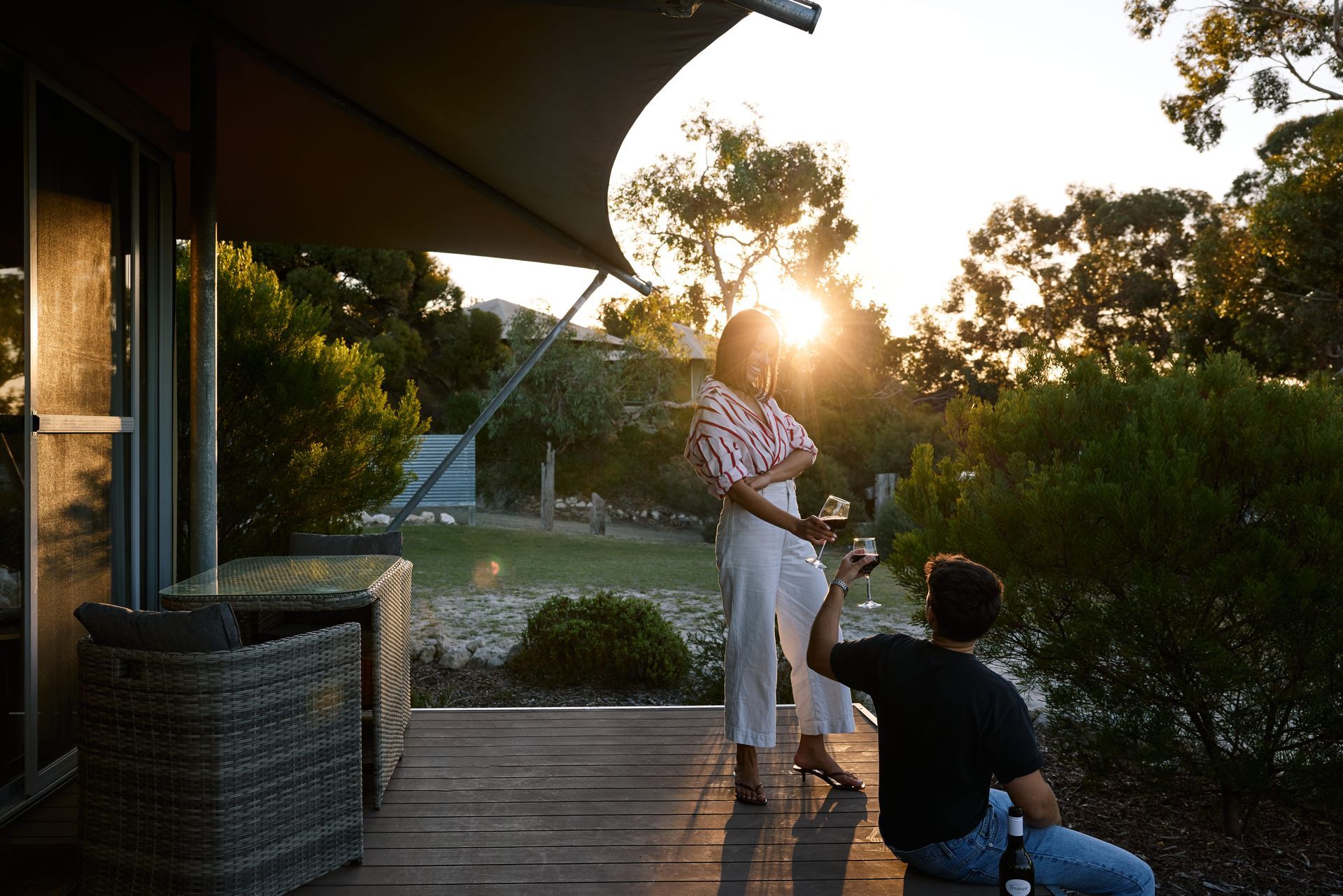 Couple on a deck toasting drinks, with sunset view, trees, and modern building.