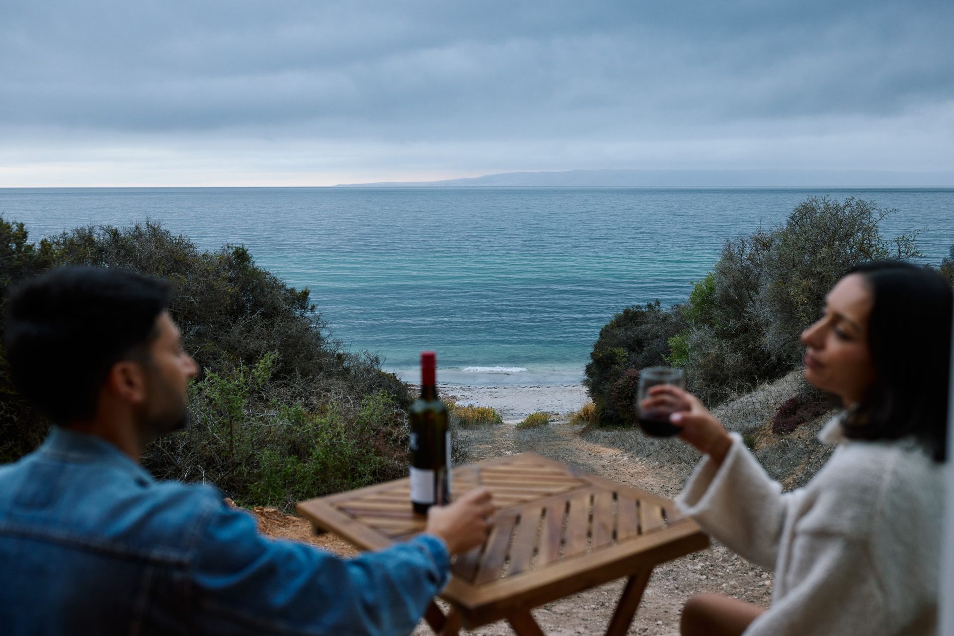Couple drinking wine, ocean view. Table with bottle and glasses. Overcast sky, shoreline in background.