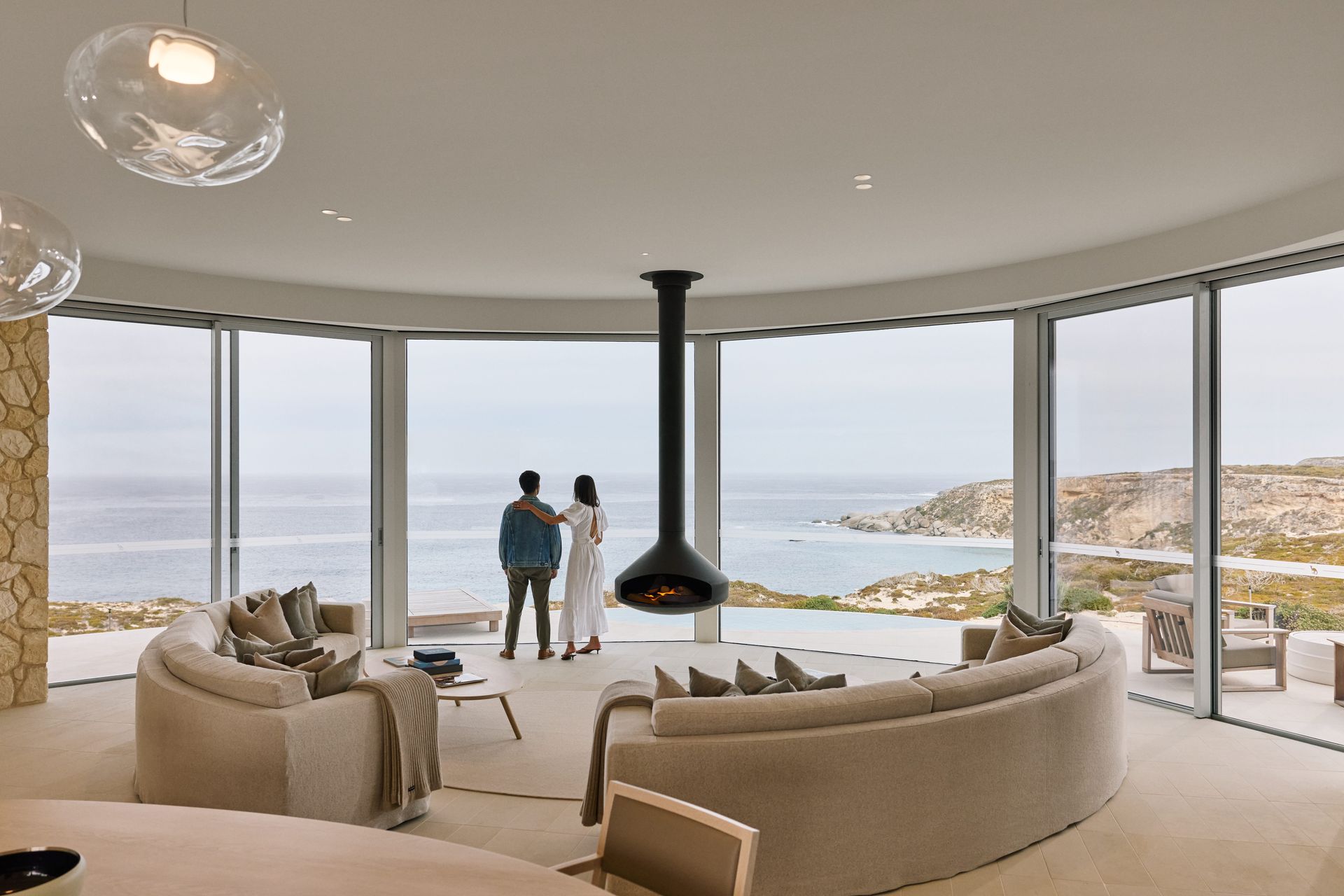 Couple standing by windows, gazing at ocean view, in modern, curved-walled living room.