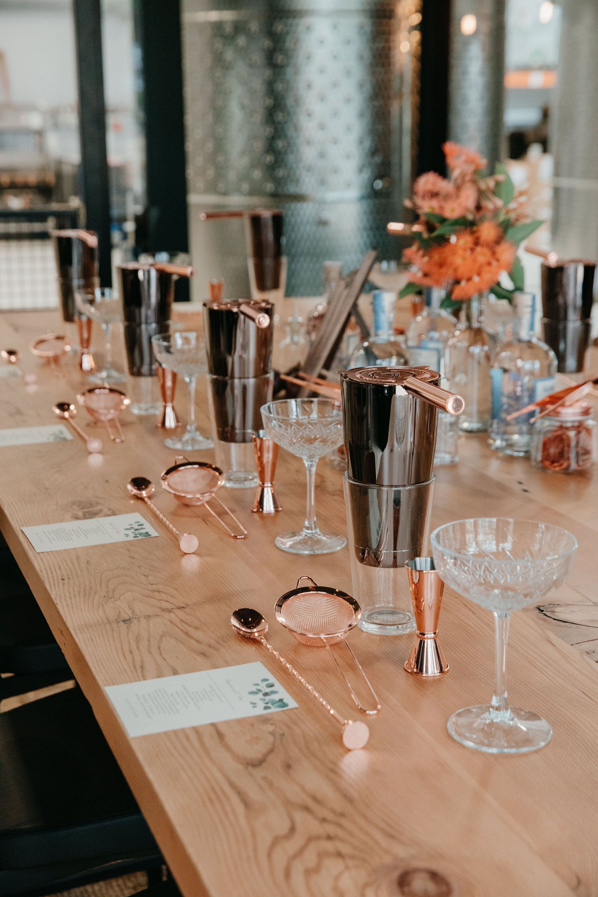 Cocktail making setup on a long wooden table with bar tools, glasses, and flower arrangement.