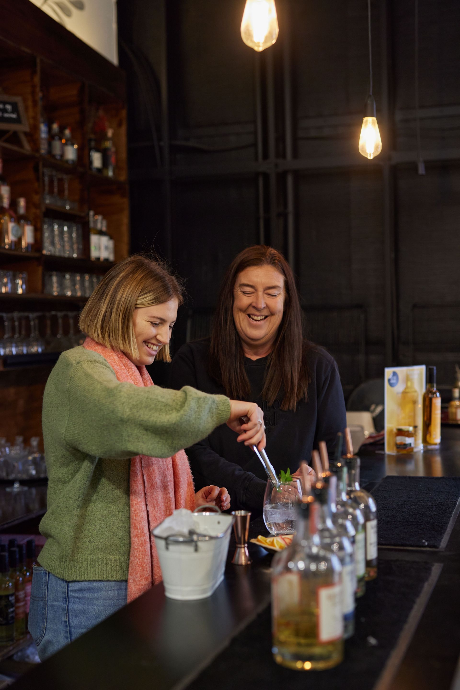 Two people laughing, mixing cocktails behind a bar, lit by hanging lights.