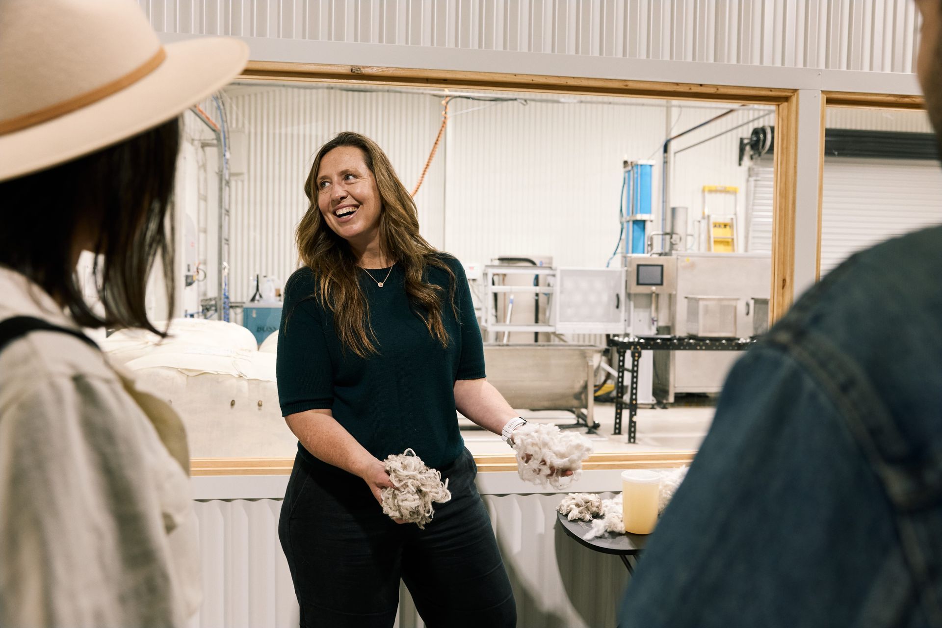 Woman in dark shirt smiles, gesturing at product samples. Two people listen; factory equipment in background.