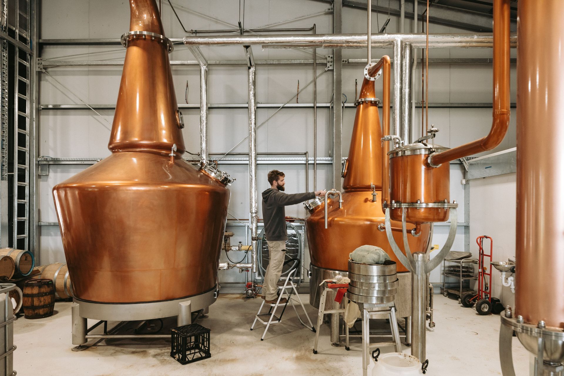 Man working on copper stills in a distillery, stainless steel equipment, light-colored interior.