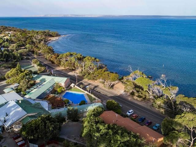Aerial view of a coastal hotel with a pool, road, and ocean; blues, greens, and browns dominate the scene.