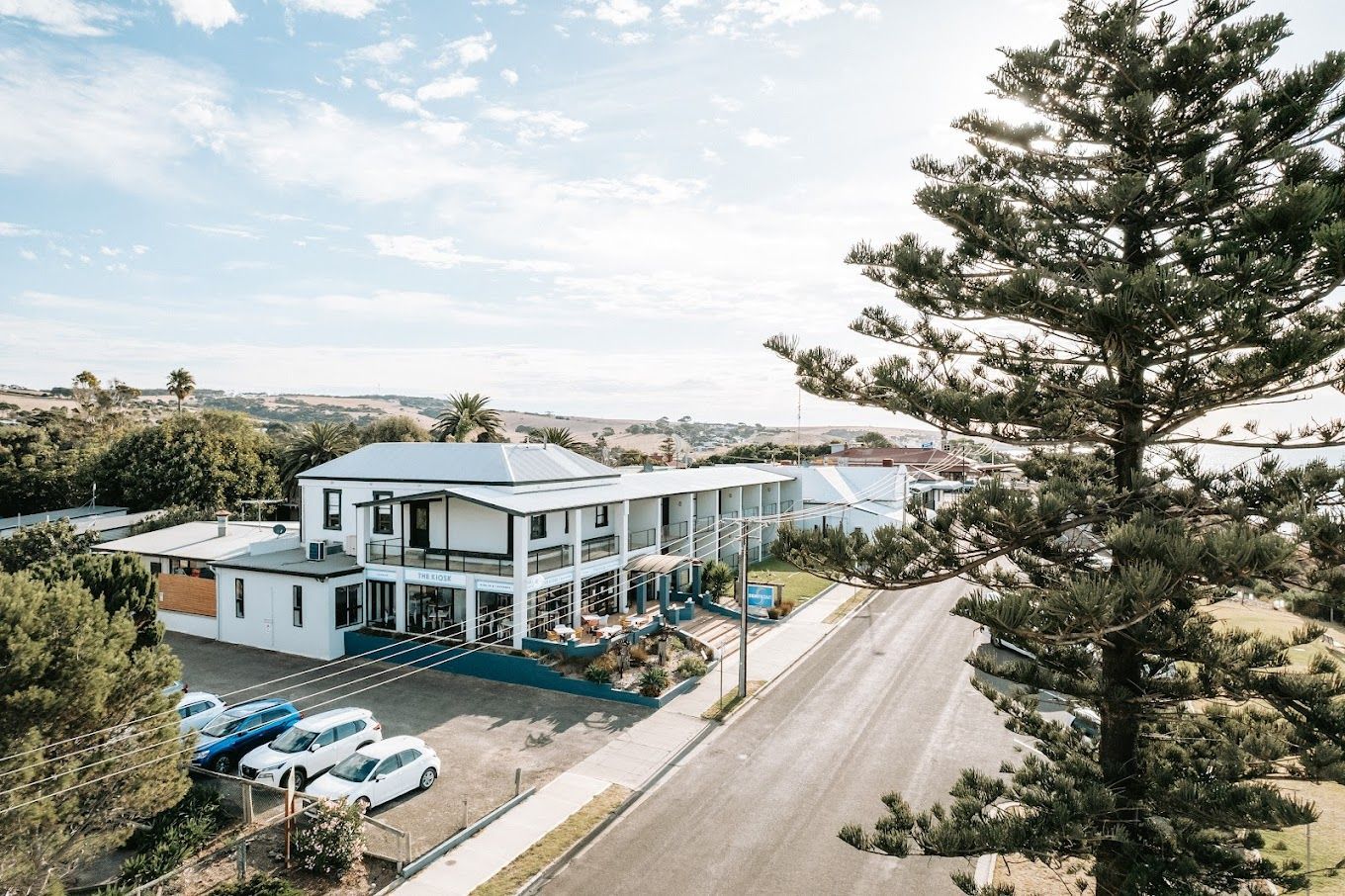 Two-story white building with blue trim, cars parked outside, tall tree, and road.