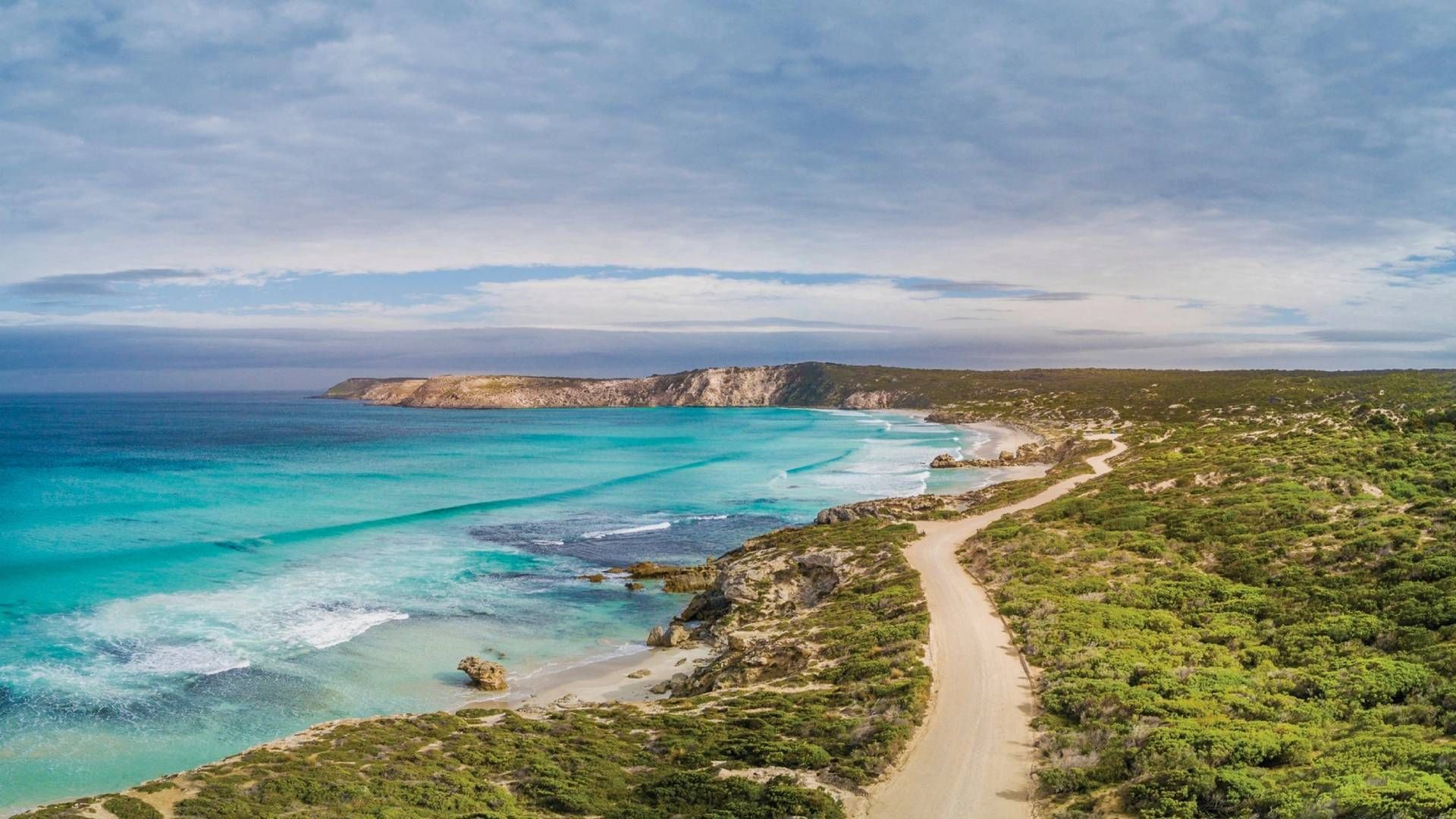 Turquoise water laps a sandy shore, a dirt road winds through green scrub, under a cloudy sky.