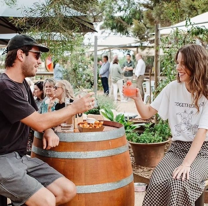 Man and woman toasting drinks, seated at a barrel table outdoors. People in background.