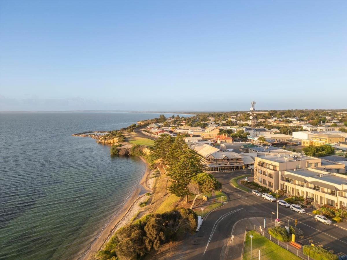 Coastal town view with buildings, road, trees, and water under a blue sky.
