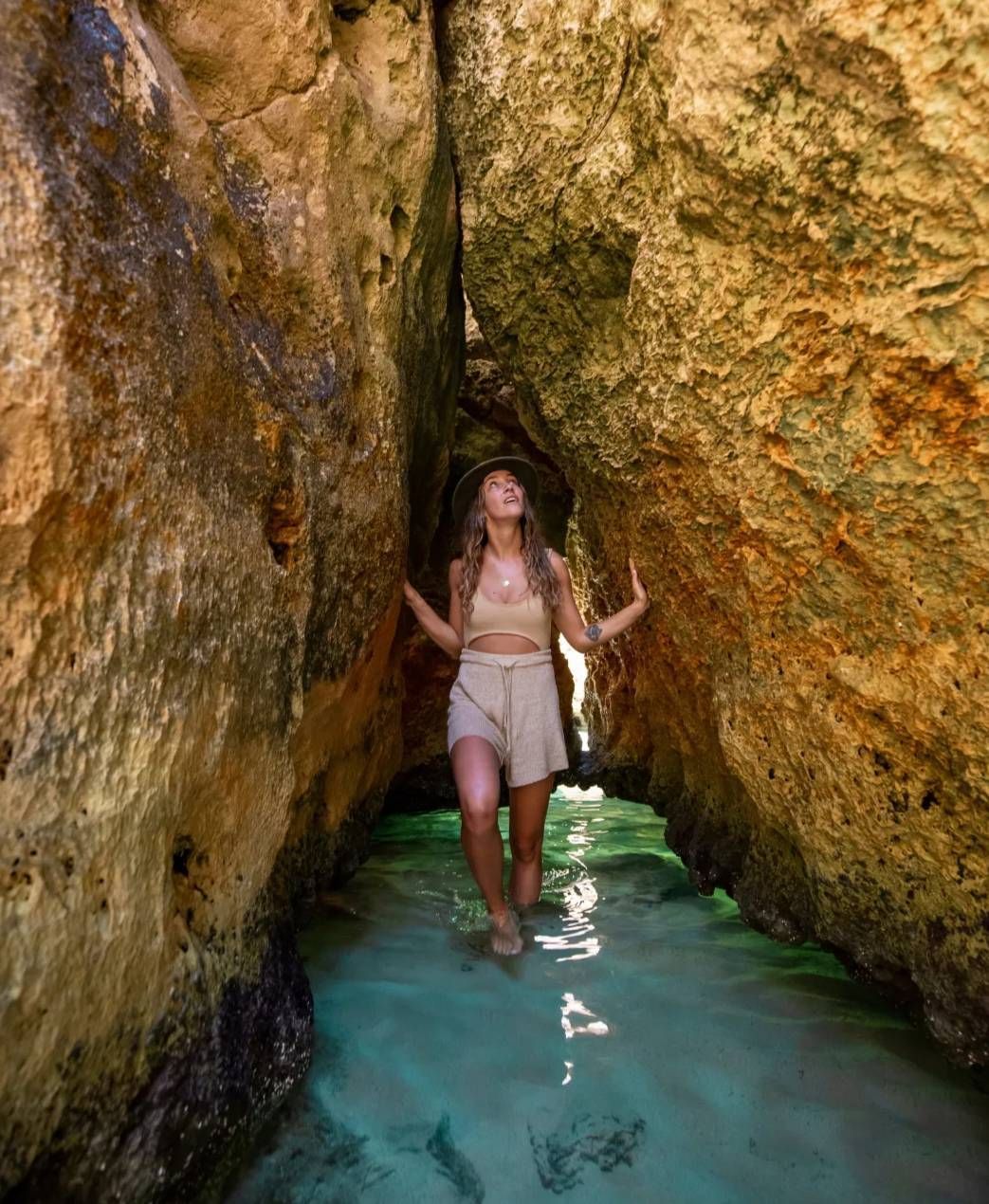 Woman wades through shallow, clear water between rock walls. Tan shorts, top, and hat.