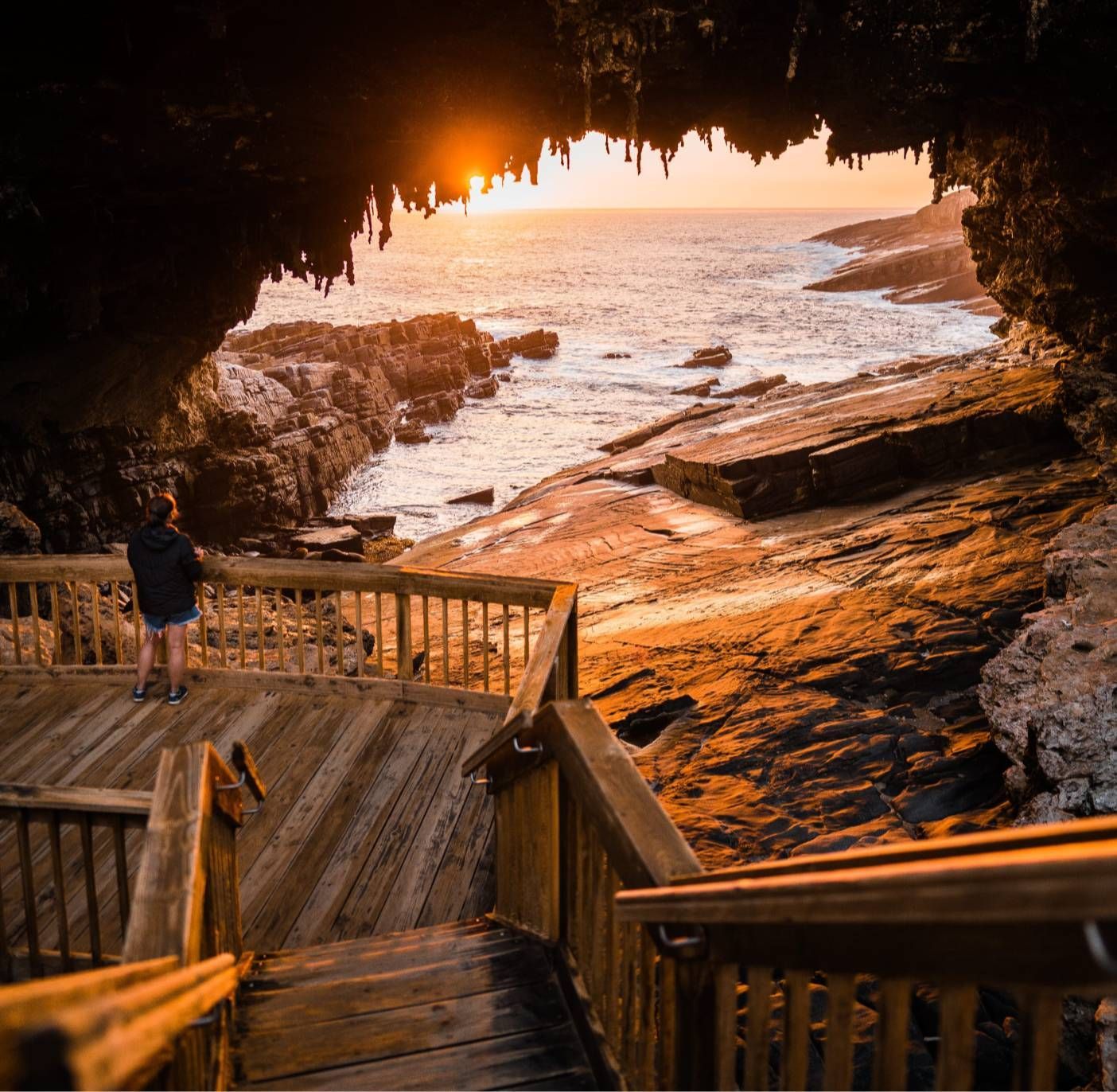 Wooden staircase leading to a coastal cave at sunset. Person looks out at the ocean. Warm golden light.