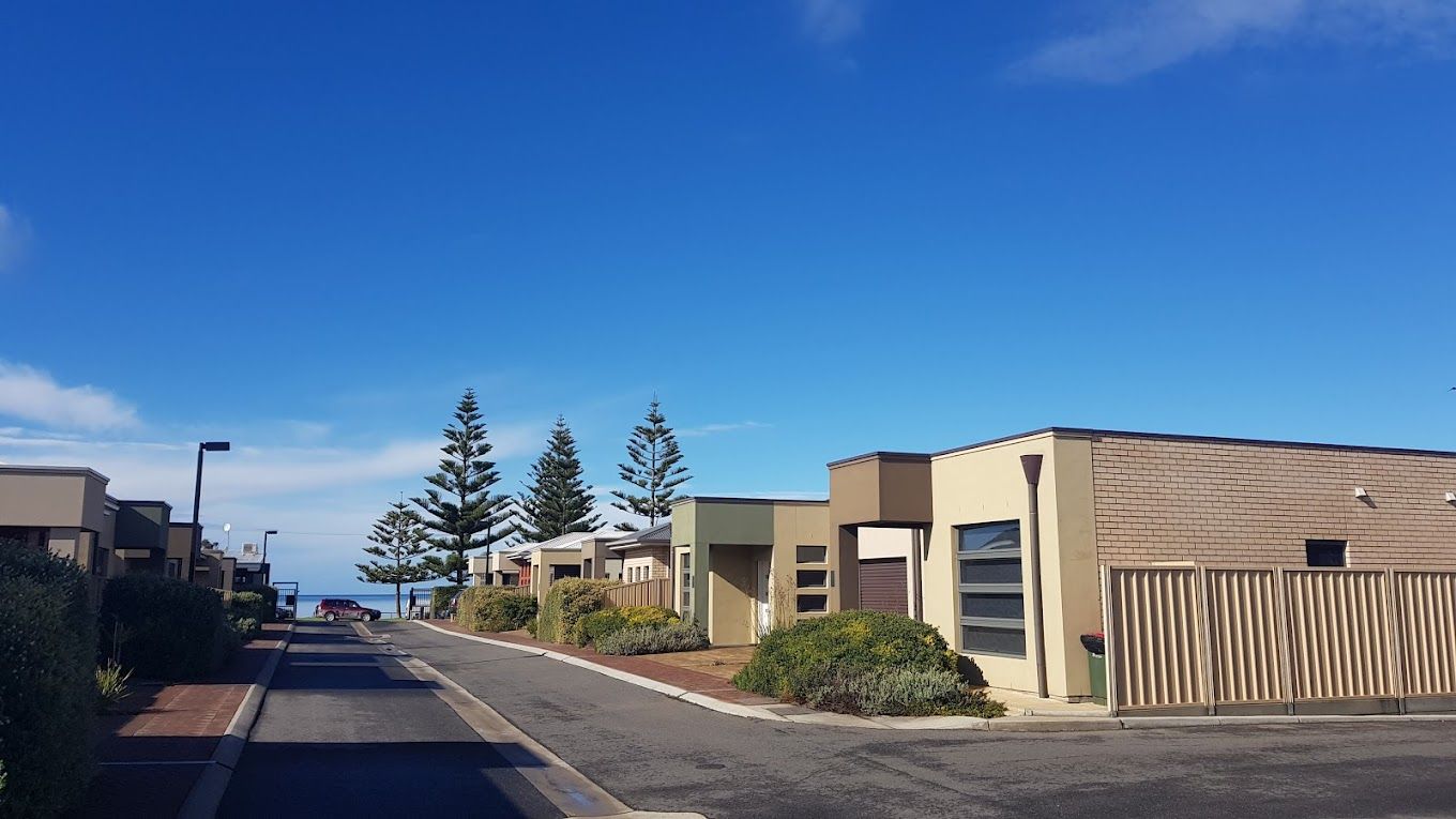 Street with houses on a sunny day, blue sky, trees, and glimpses of the ocean.