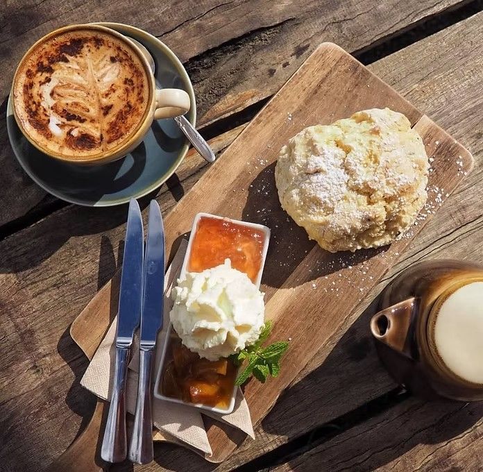 Latte, scone with jam and cream, teapot on wooden board. Outdoor setting, sunny.