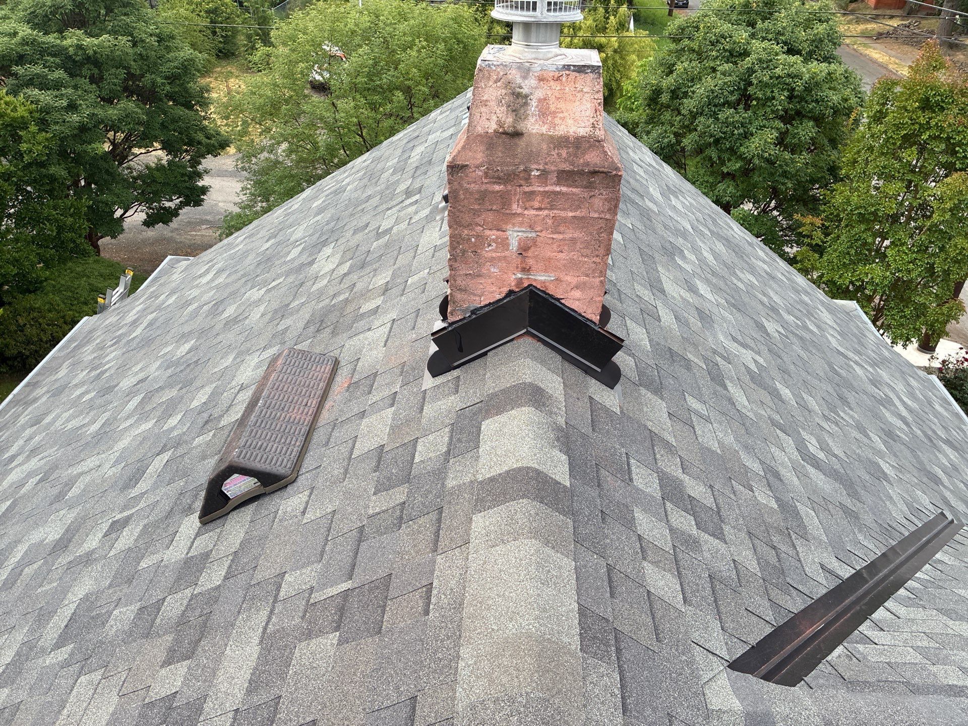 Aerial view of a gray shingled roof with a red brick chimney, surrounded by green trees.