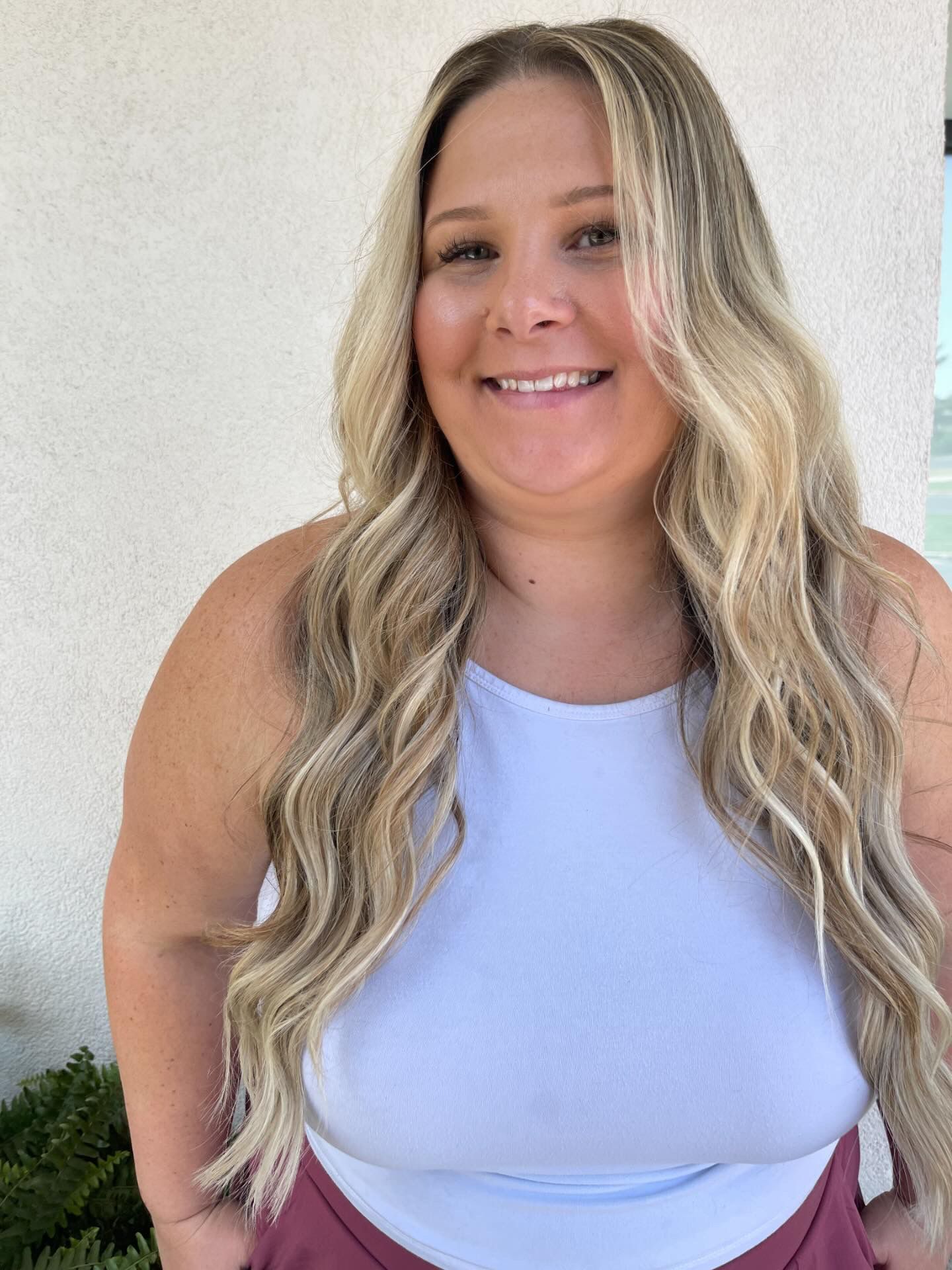 A woman with long blonde hair is wearing a white tank top and smiling.
