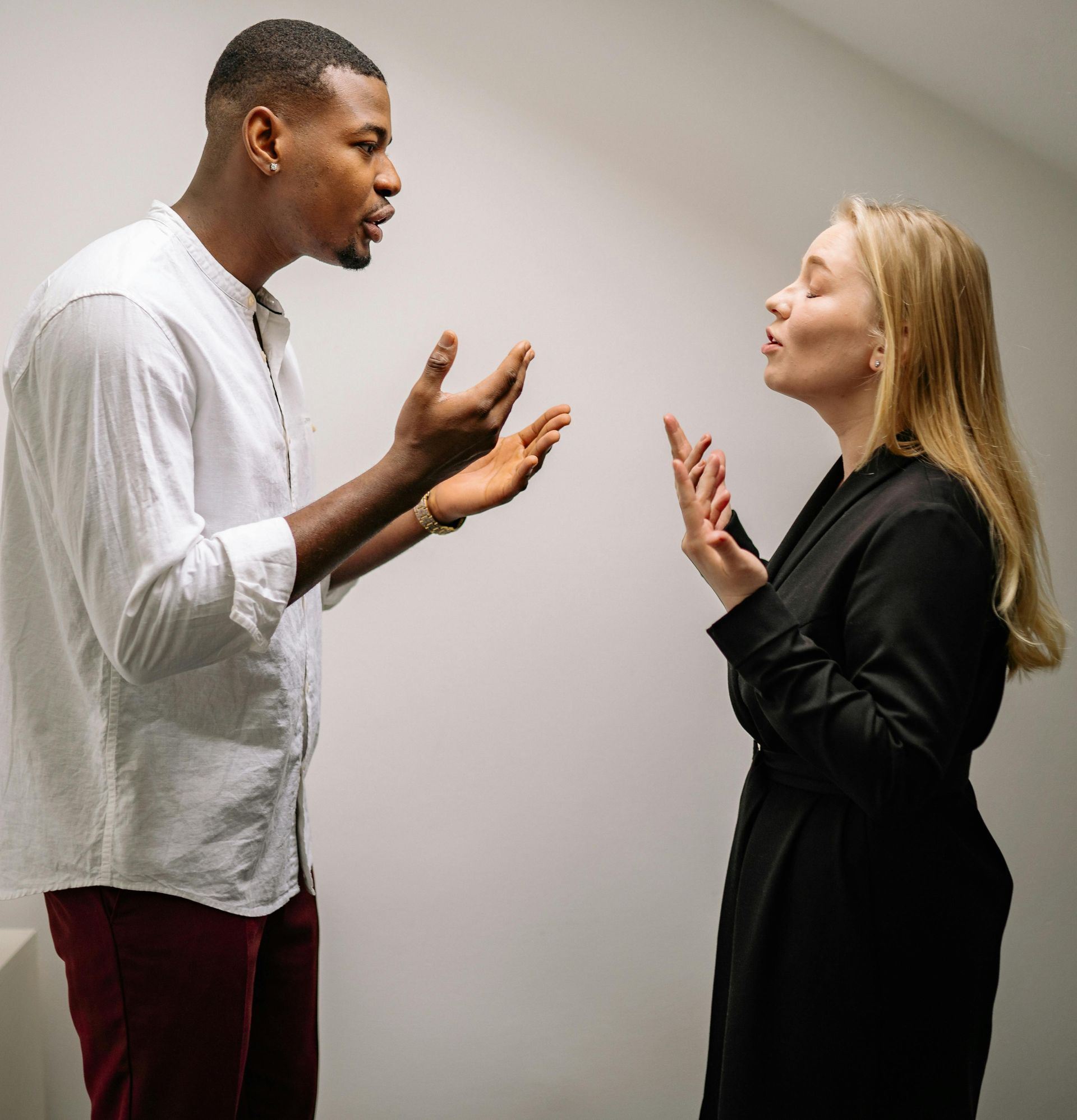 A man and a woman argue; they gesture with hands in a room with a white wall.