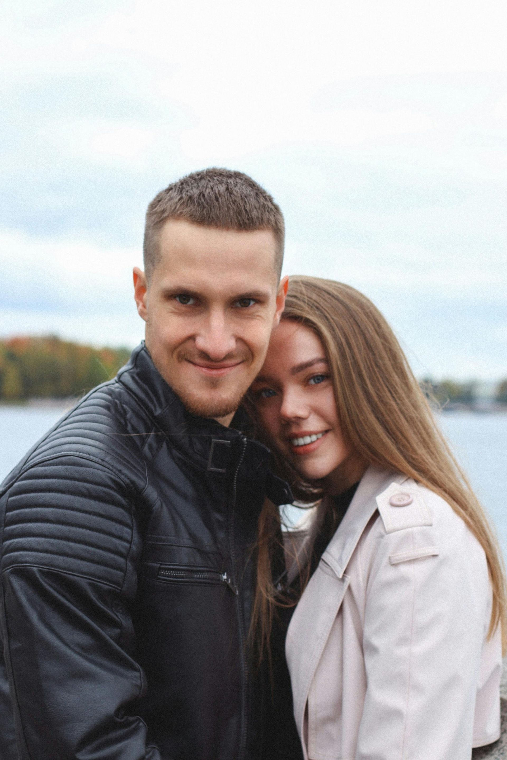 A couple smiling and hugging outdoors by the water, with cloudy sky in the background.