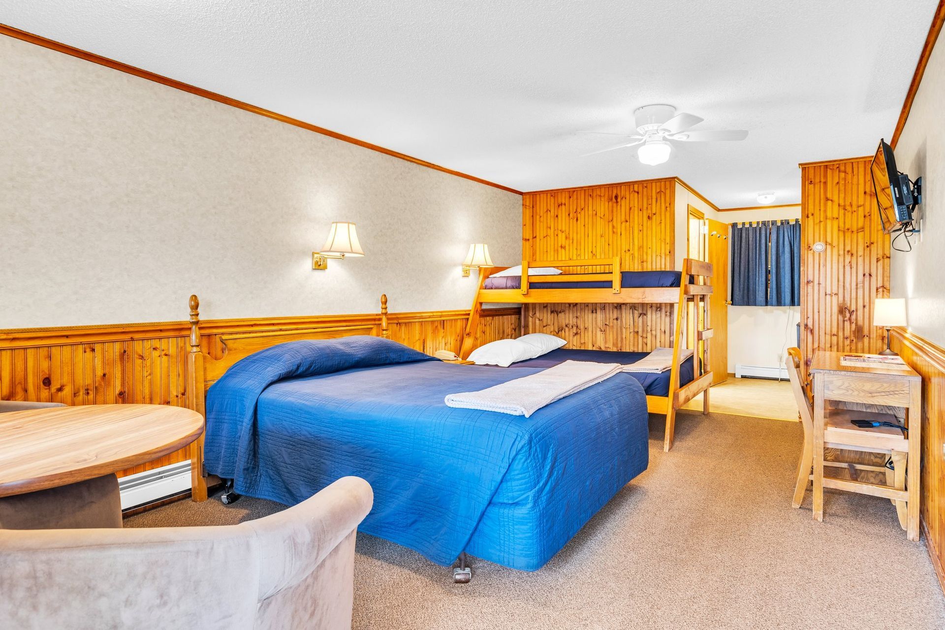 Hotel room with a queen bed, bunk beds, wood paneling, and a small table.