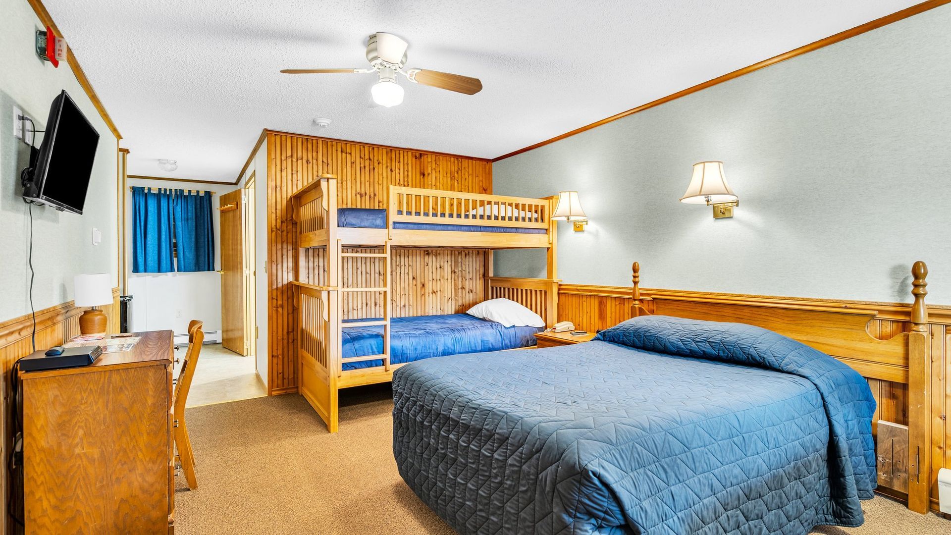 Hotel room with double bed, bunk beds, desk, and TV. Walls are paneled with wood.