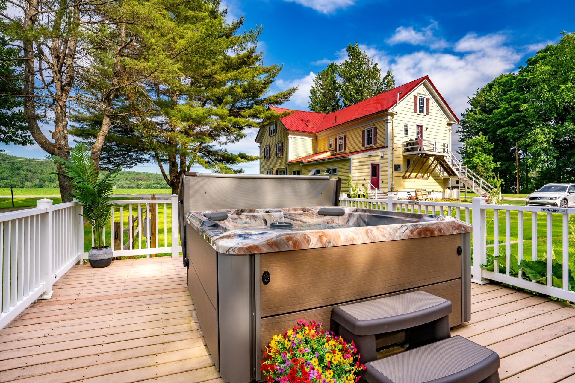 Hot tub on a deck overlooking a rural landscape with a yellow house in the background.