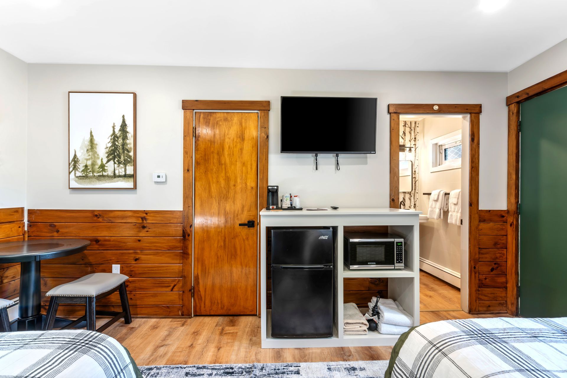 A small motel room with a mini-fridge, microwave, TV, and two beds. Wooden accents and a forest painting.