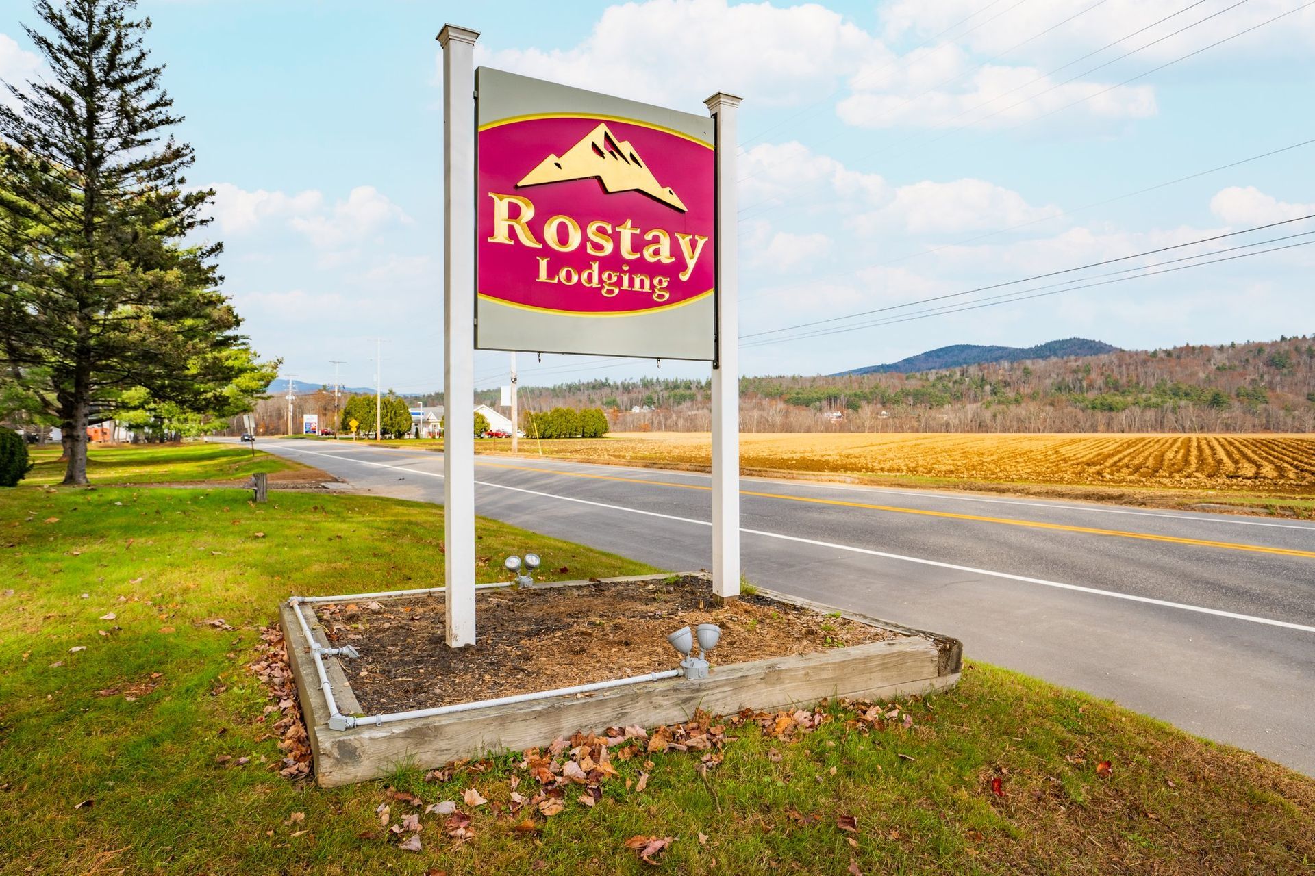 Sign for Rostay Lodging with a mountain logo by a road with fields and trees.