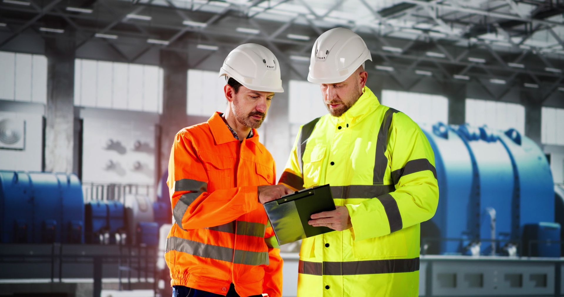Two construction workers are looking at a clipboard in a factory. Two construction workers are looking at a clipboard in a factory.