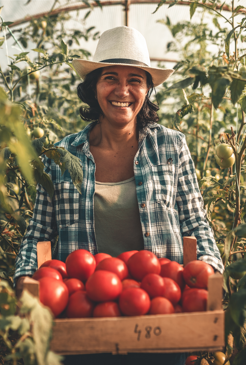 Woman holding a wooden crate overflowing with red tomatoes, smiling in a greenhouse.