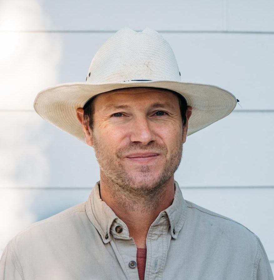 Man in a straw hat smiles, wearing a light-colored button-down shirt against a white paneled wall.
