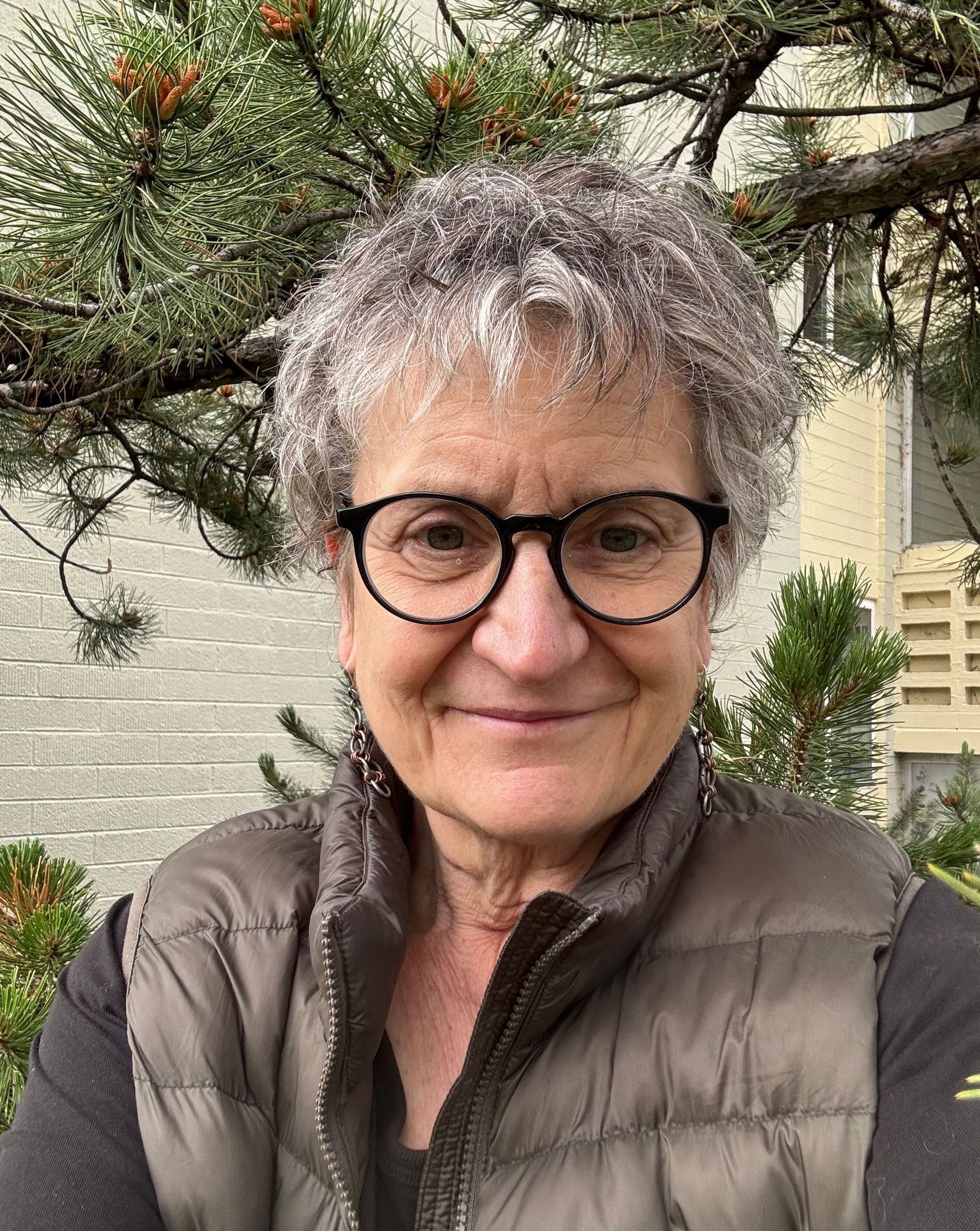 Woman with glasses, short gray hair, and vest smiles outside near greenery.