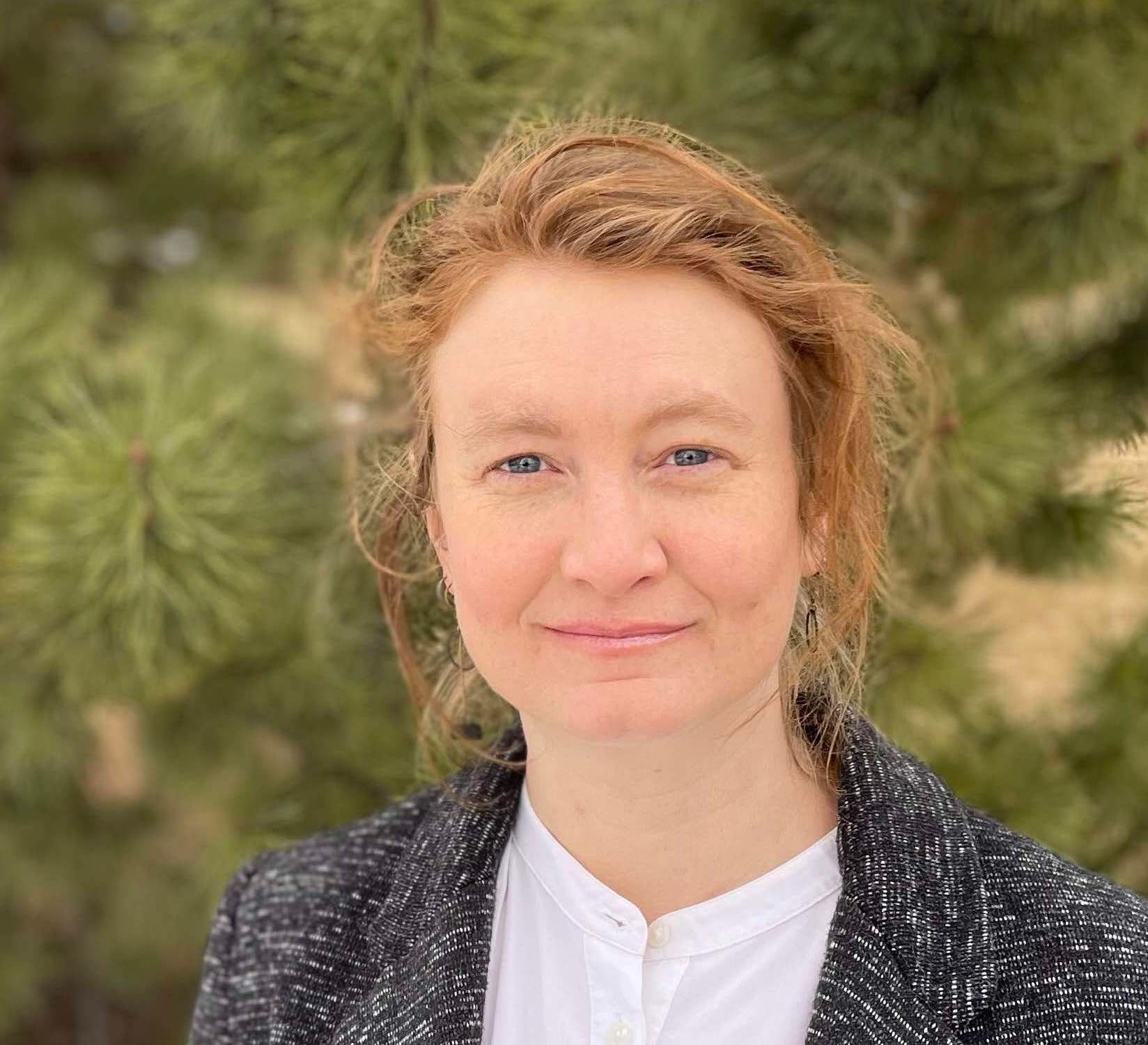 Woman with red hair, smiling, wearing a white shirt and gray blazer, in front of evergreen trees.