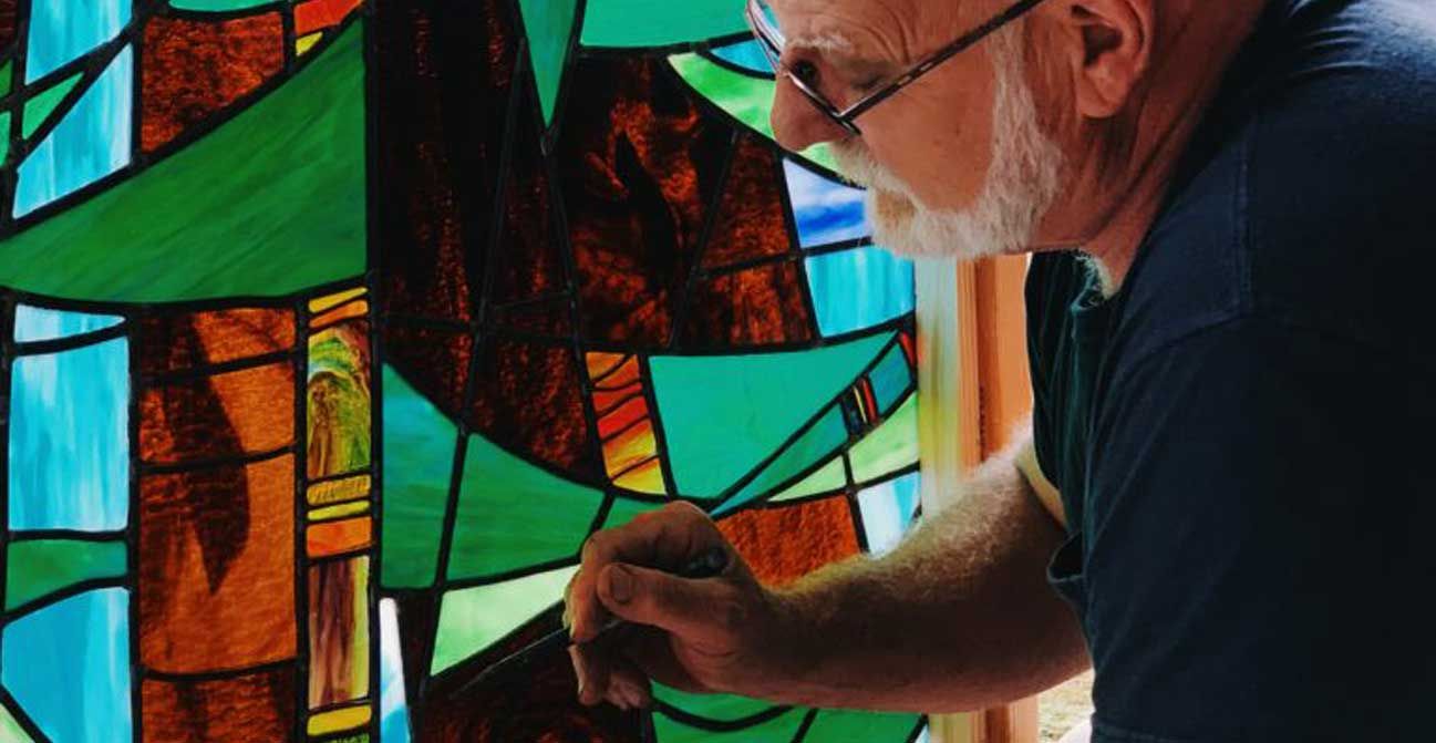 Man working on stained glass with colorful panes, wearing glasses and a dark shirt.