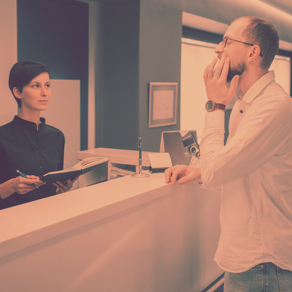 Woman at desk with pen, facing man holding hand to face. Office setting, warm colors.