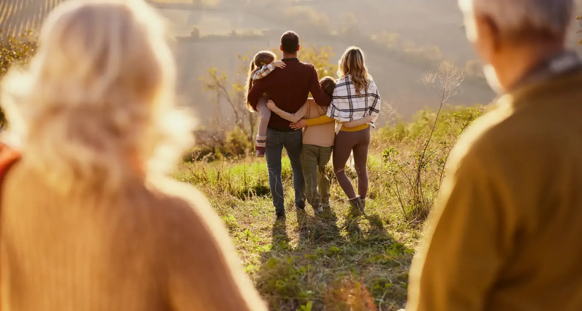 Family of five walking away, view from behind of older couple. Sunlight, open field.