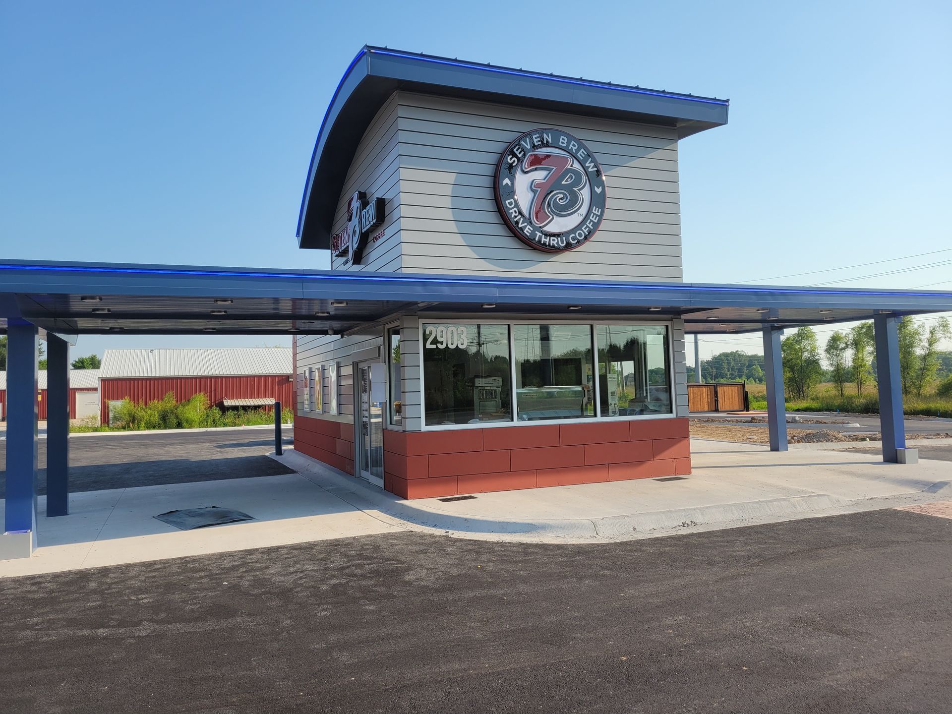 A modern drive-thru building with a gray, brick-patterned facade, red base, and a blue canopy under a clear blue sky.