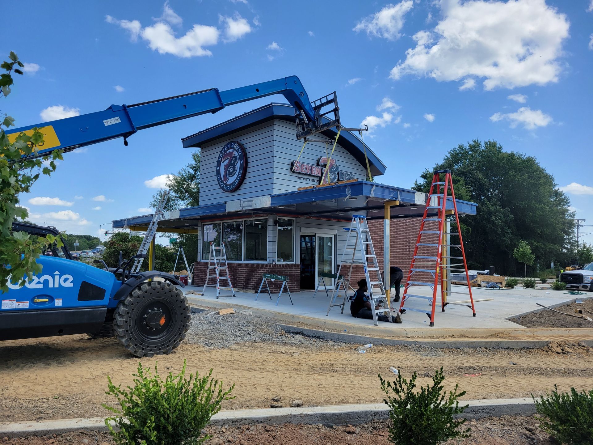 A blue Genie lift raises workers toward the roof of a building under construction, with ladders and gravel surroundings.