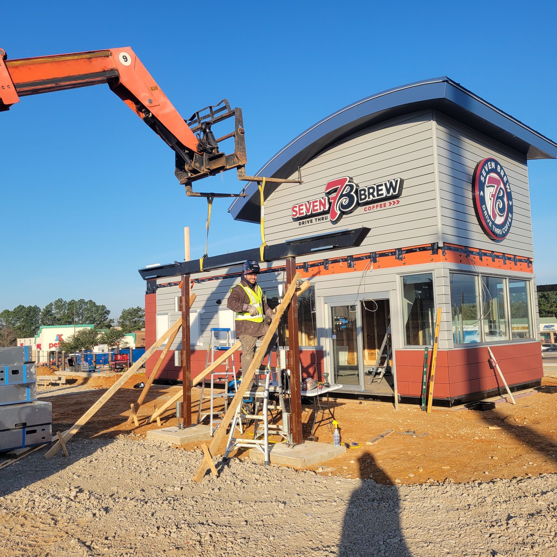 A worker in a high-visibility vest stands on a ladder next to a modern Seven Brew coffee building under construction.