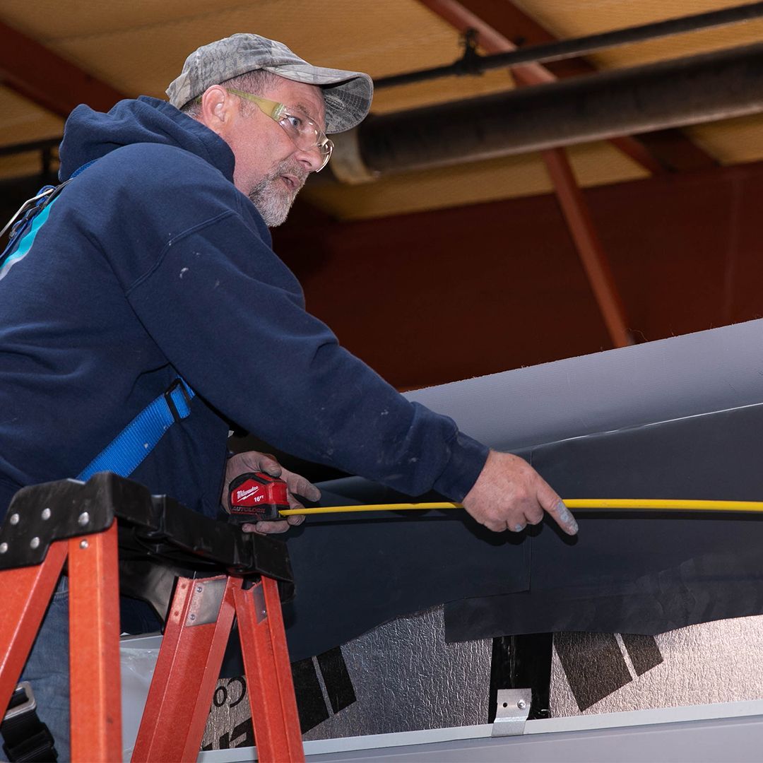 A worker in a blue hoodie and camo cap stands on an orange ladder, measuring a dark material with a yellow tape measure.