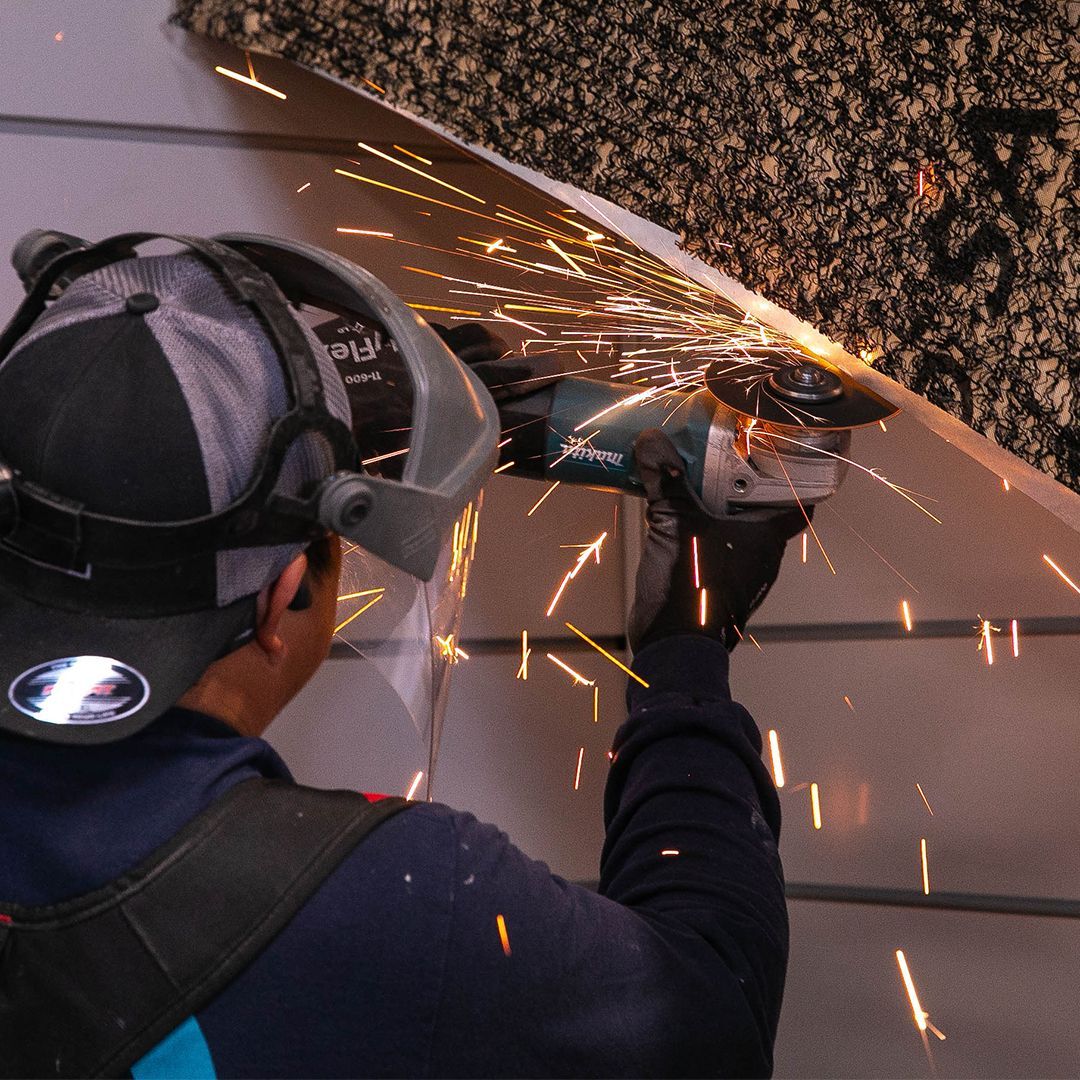 A worker wears a face shield and gloves while using an angle grinder to cut metal, creating sparks.