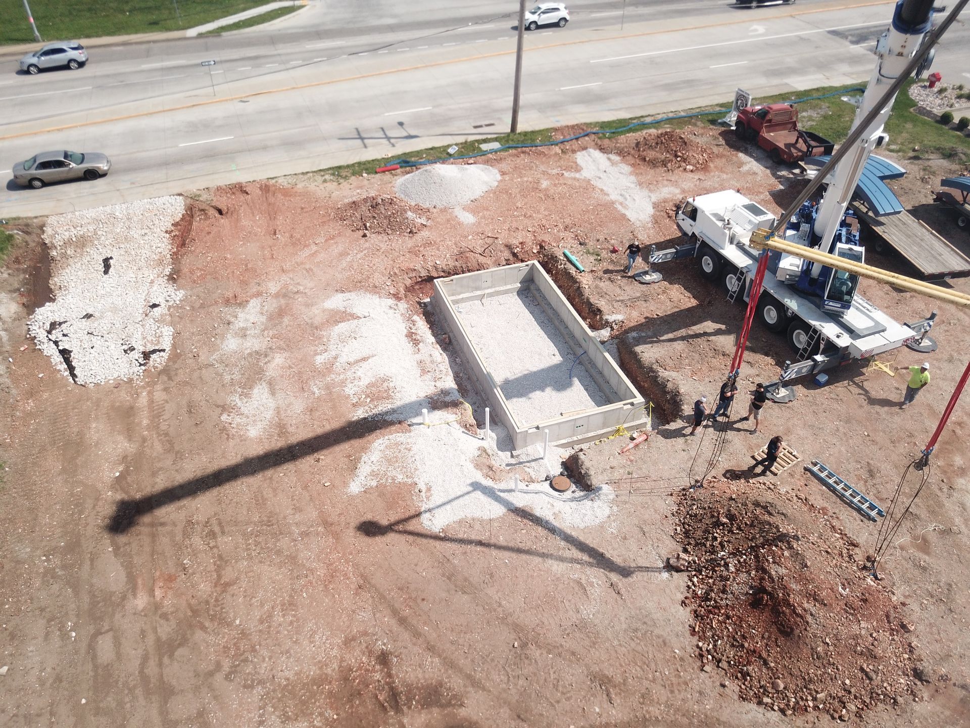 An aerial view of a construction site showing a crane installing a rectangular swimming pool shell in a dirt lot.