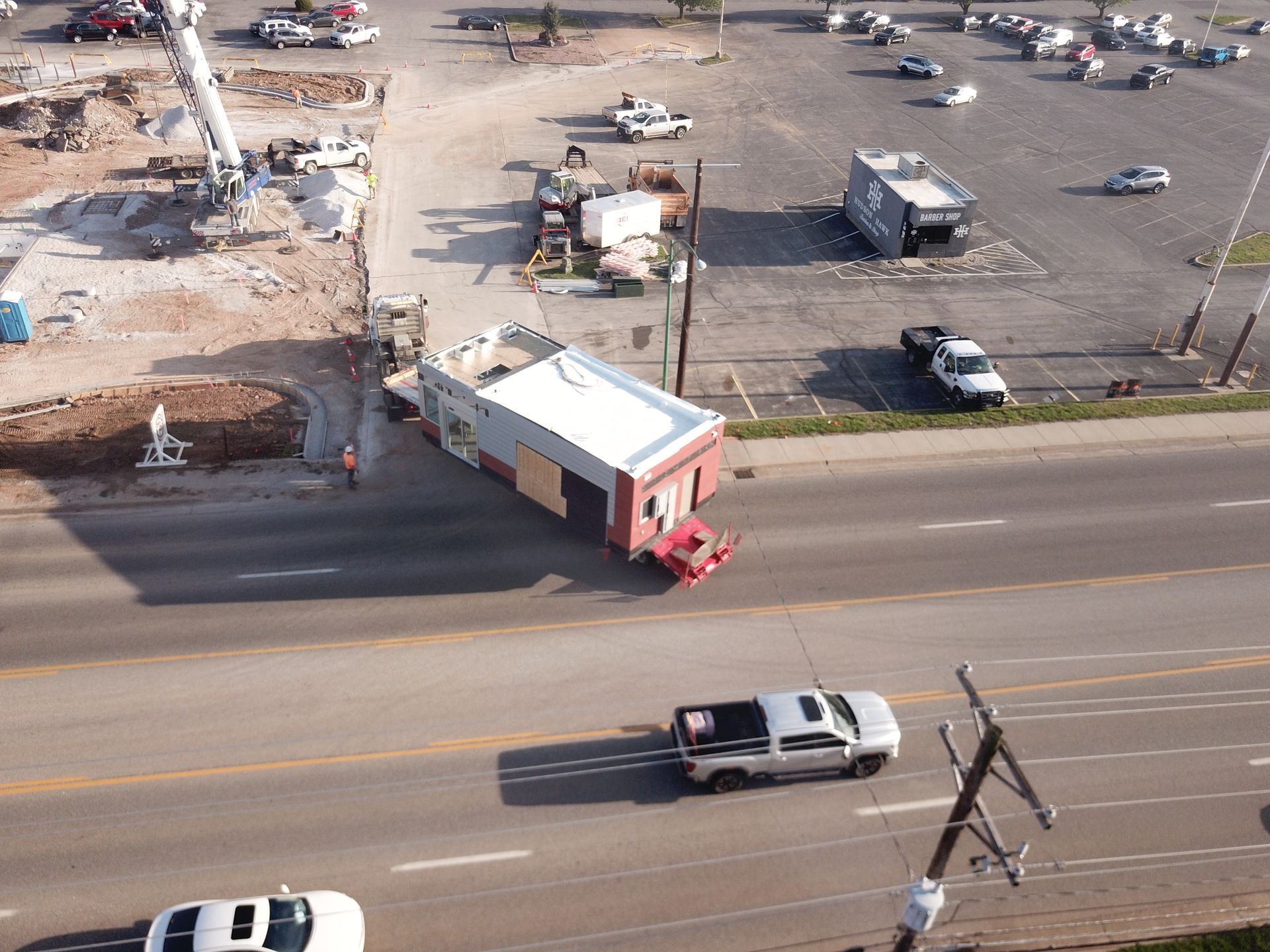 An aerial view shows a modular building being transported by truck along a paved road near a construction site and parking.