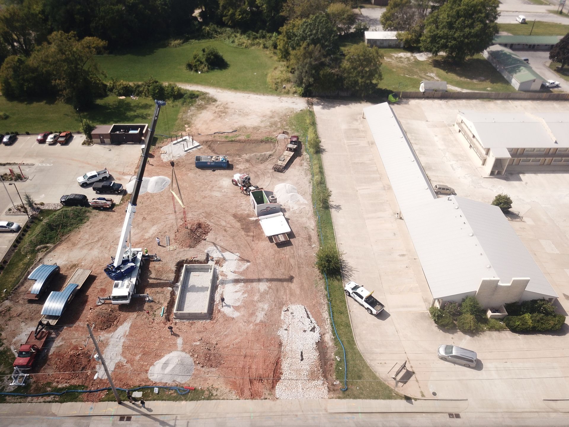 Aerial view of a construction site with a crane, a rectangular pool shell, trucks, and equipment on a dirt lot.
