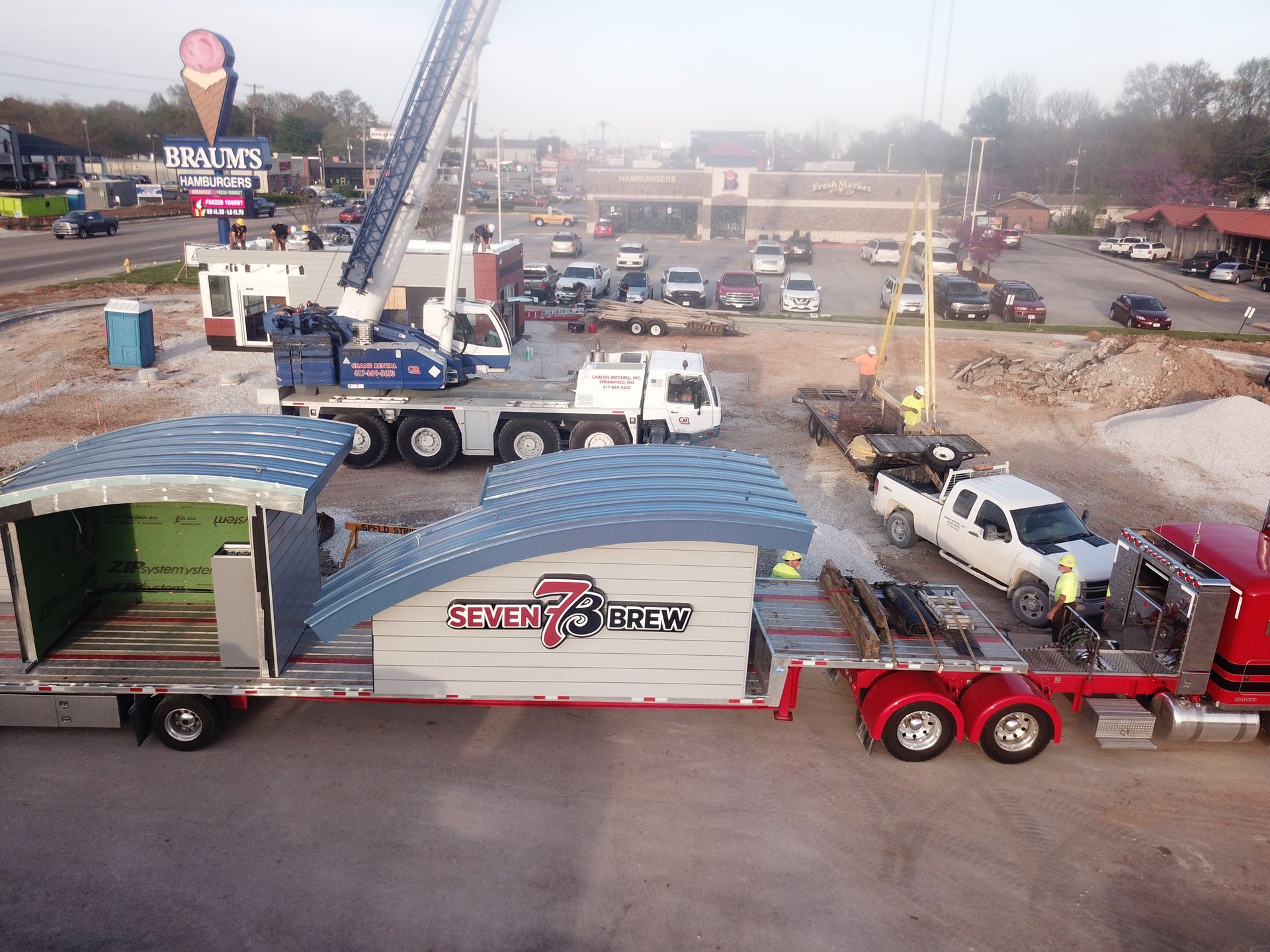 A crane lifts construction sections onto a red flatbed truck at a site near a retail business with an ice cream sign.