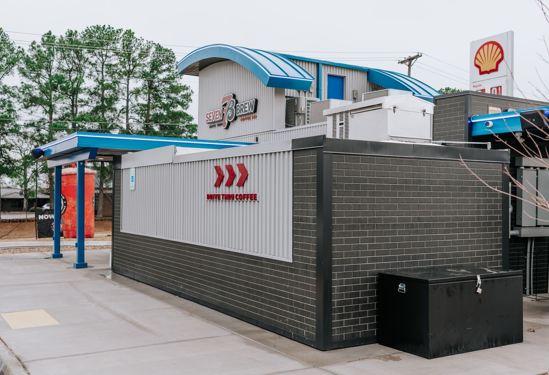 A modern, industrial-style outdoor structure with a blue curved roof, grey metal siding, and dark brick walls near a Shell.