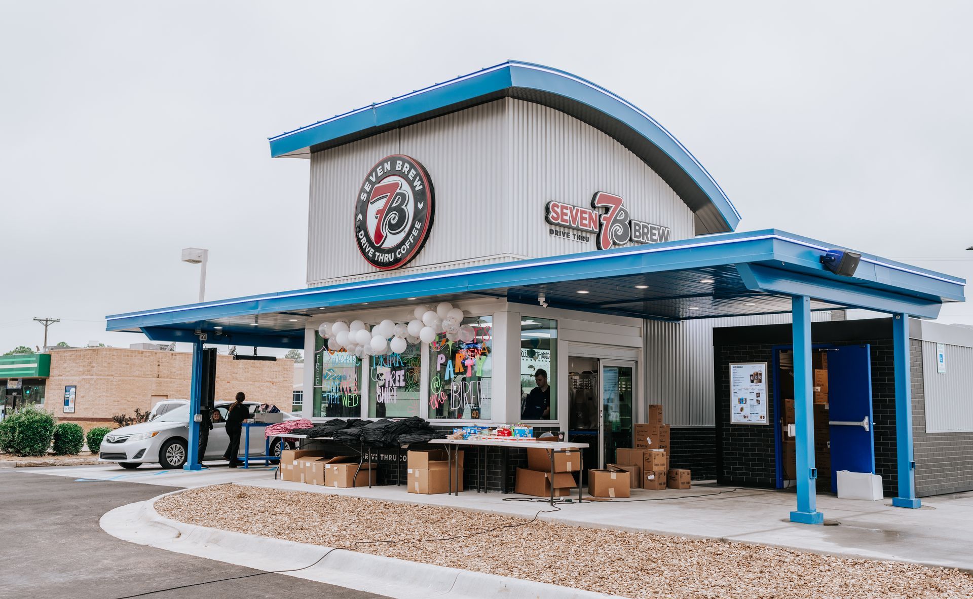 A white car wash building with a blue metal roof and trim, decorated with white balloons, under a bright, overcast sky.