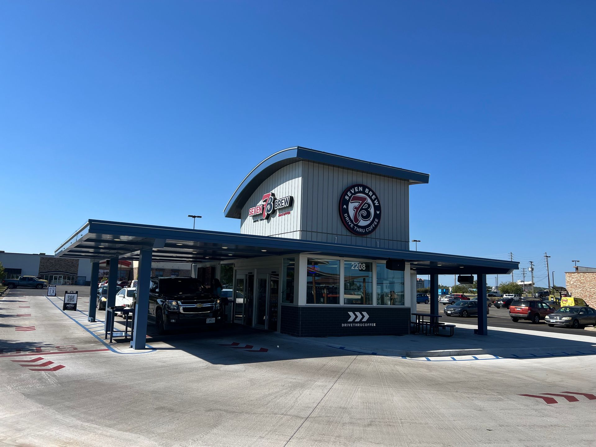A modern, blue-roofed 7 Brew drive-thru coffee stand under a bright blue sky, with a car approaching the service window.