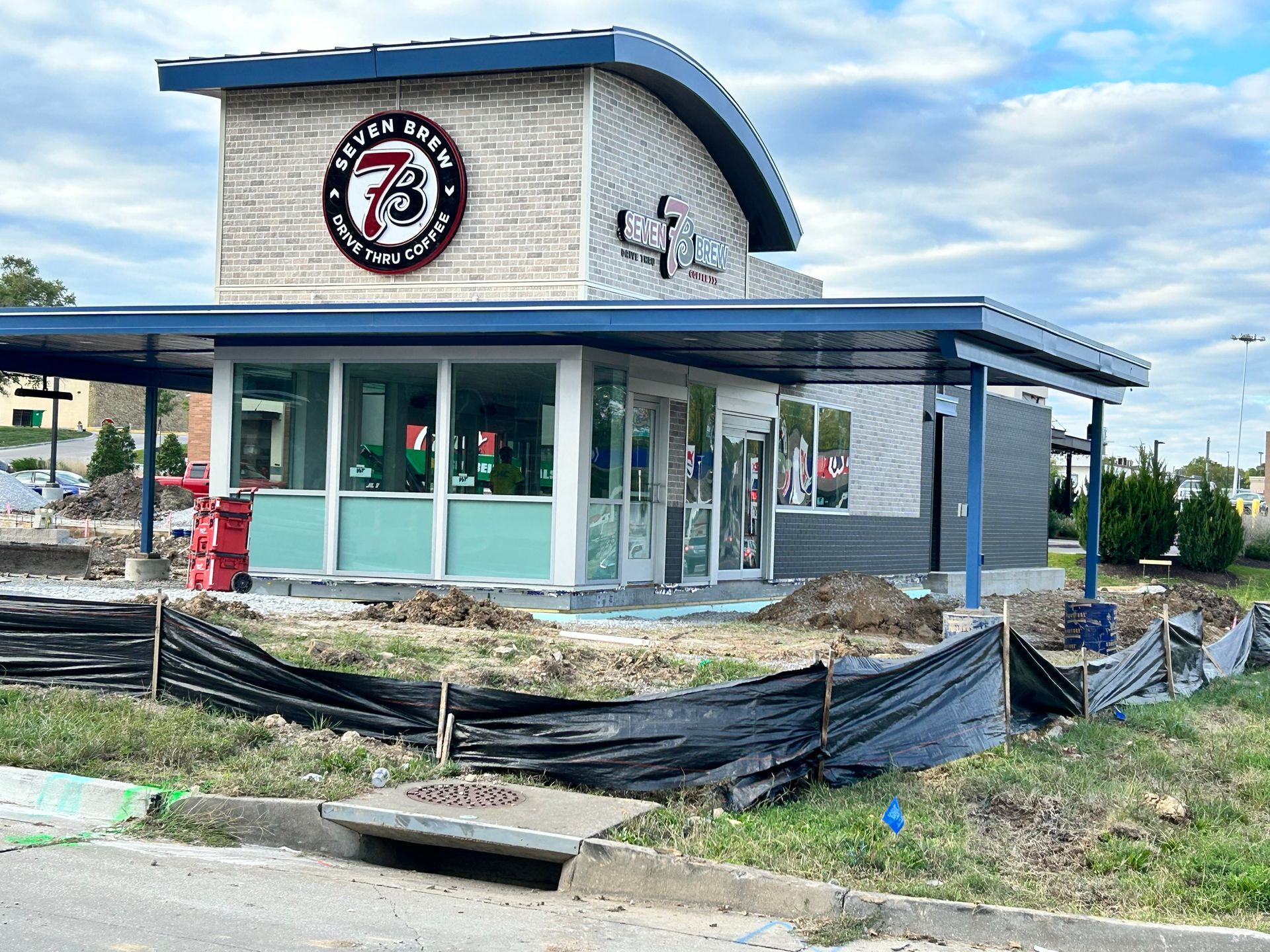 A modern Seven Brew drive-thru coffee stand under construction with a tan stone facade, blue roof, and black fencing.
