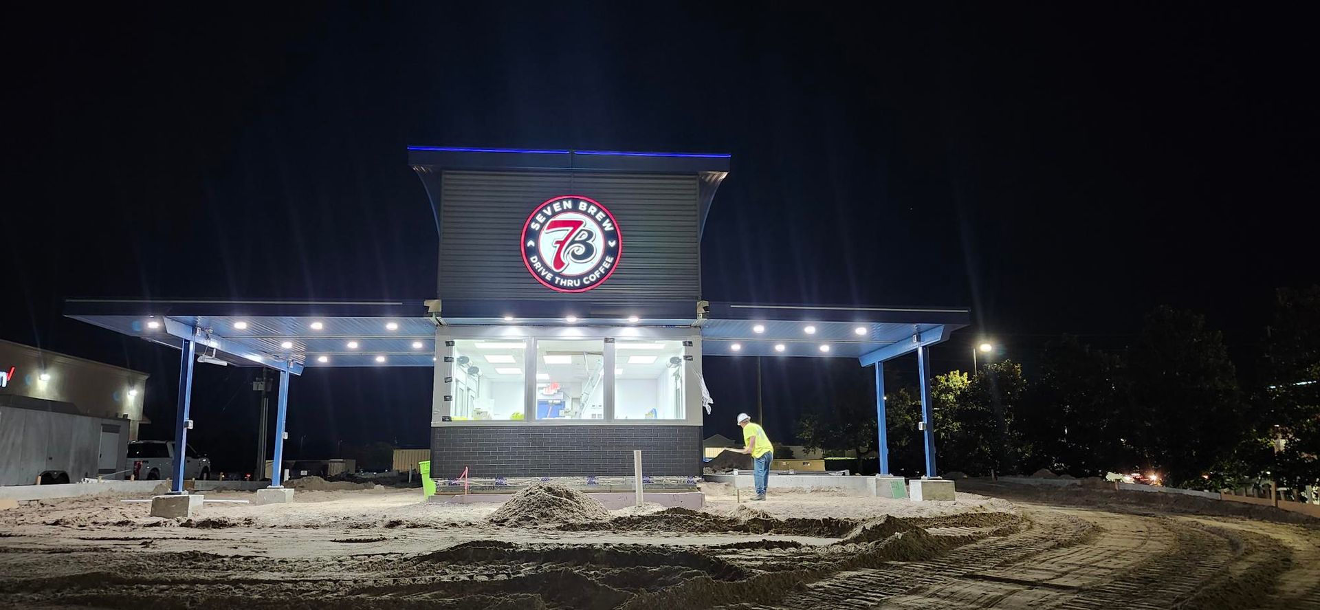 A modern, illuminated gas station at night, featuring a prominent circular logo and a worker standing in the lot.