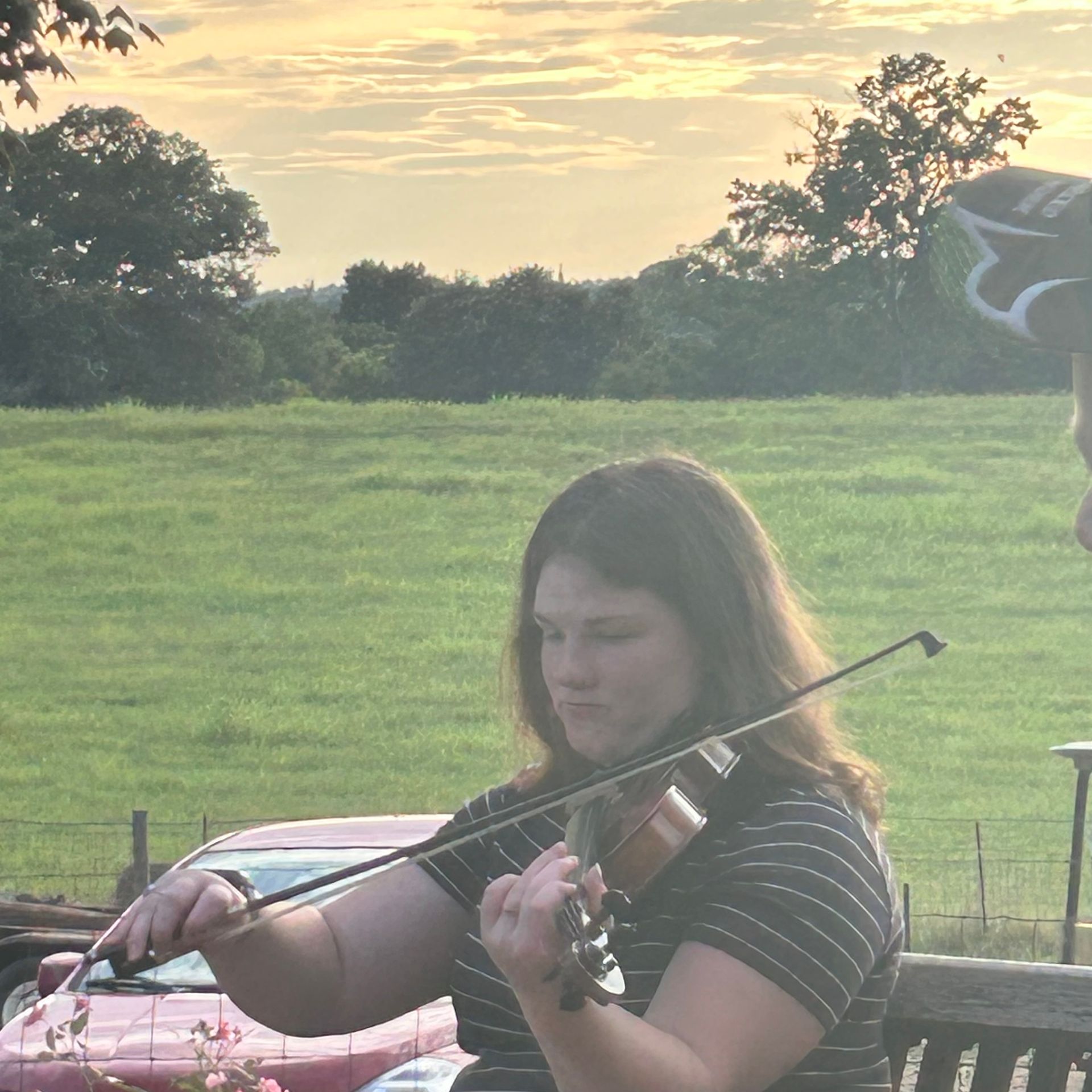 Woman playing a violin outdoors, with a field in the background during sunset.