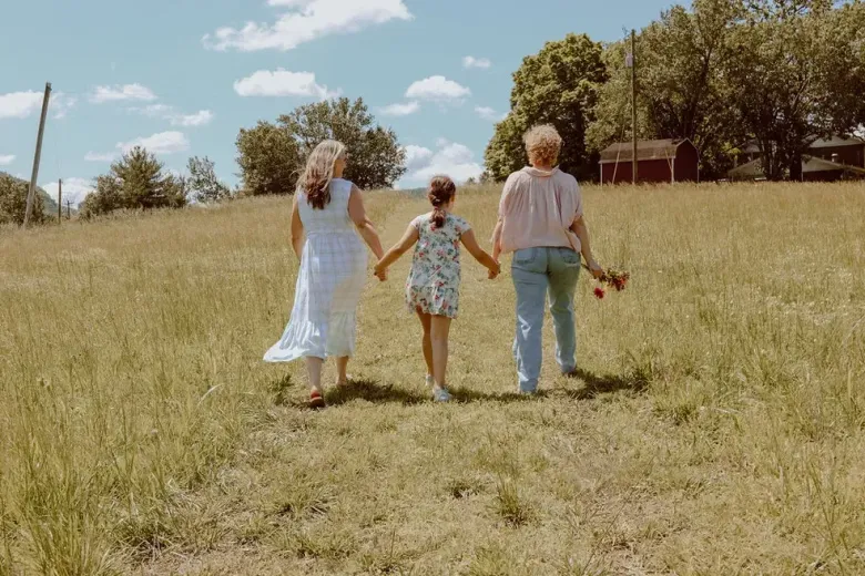 Three women and a little girl are walking through a grassy field holding hands.