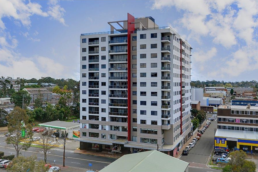 A tall white apartment building with a red vertical accent, situated on a corner in a suburban area with a nearby gas station.